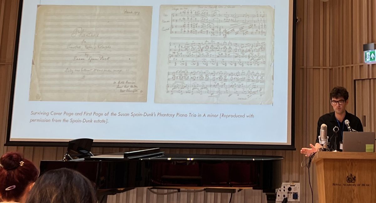 Matthew Madeley presenting to an audience at the Royal Academy of Music, with a slide showing parts of the manuscript of Phantasy Piano Trio in A minor by Susan Spain-Dunk